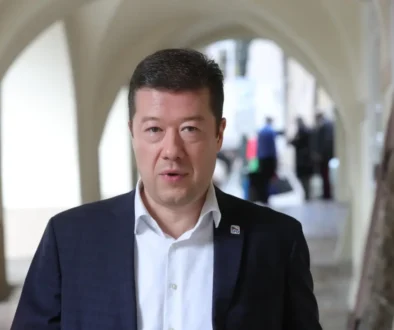 Man in a dark suit and white shirt walks toward the camera in a bright arched hallway; pin on lapel flourish act as SPD badge.