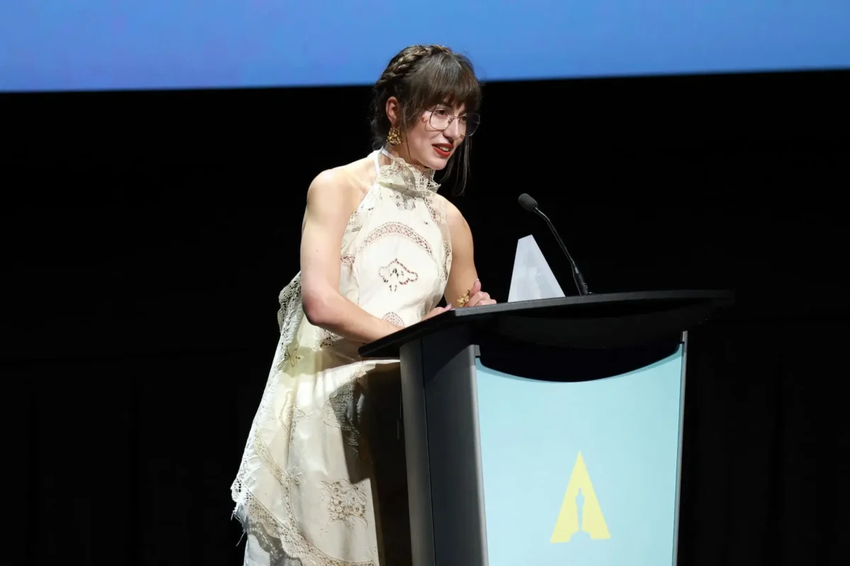 Woman in a cream lace halter dress speaks at a podium on a dark stage, with a microphone nearby and a blue panel featuring a yellow emblem on the front.