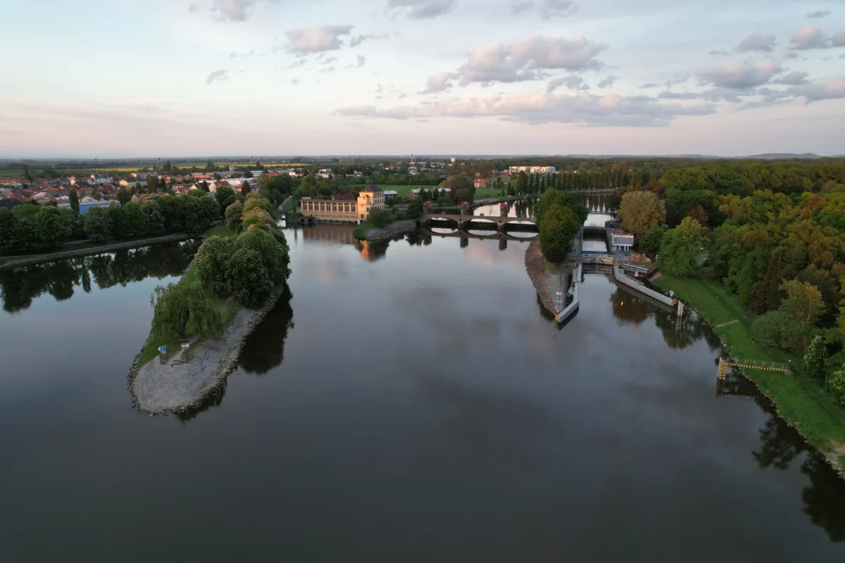 Aerial view of a calm river winding through a town, with a stone-arched bridge and a yellow riverside building, surrounded by green trees.