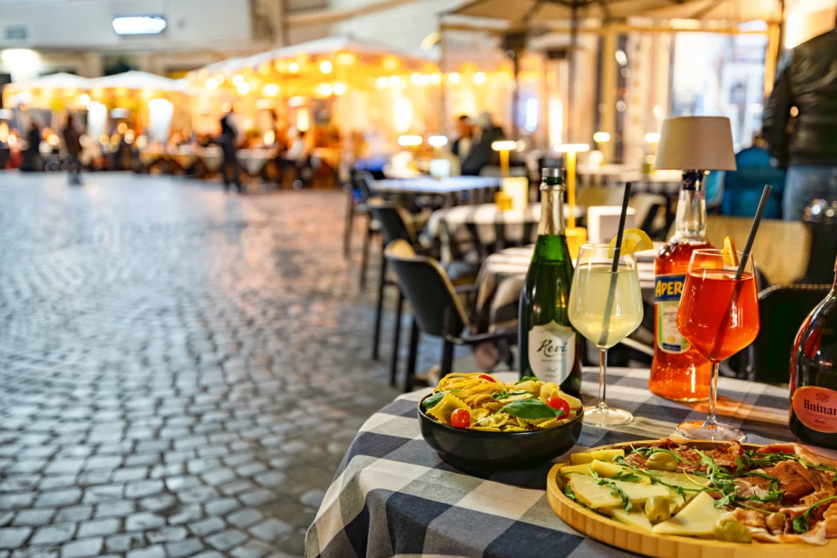 shutterstock_2739472859 1 | TVGURU.cz Outdoor night dining scene with a checkered tablecloth, drinks, and a cheese platter in the foreground; warm market lights in the background.