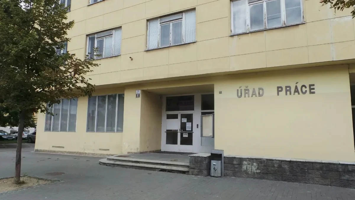 Yellow government building with the sign ÚRAD PRÁCE and glass doors at the entrance; a tree is on the left and parked cars in the background.