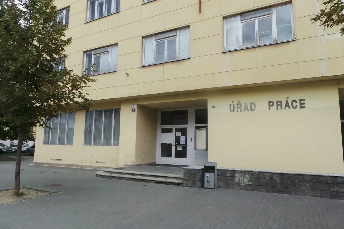 Yellow government building with the sign ÚRAD PRÁCE and glass doors at the entrance; a tree is on the left and parked cars in the background.