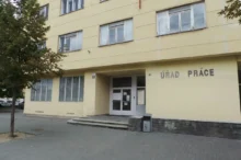 Yellow government building with the sign ÚRAD PRÁCE and glass doors at the entrance; a tree is on the left and parked cars in the background.