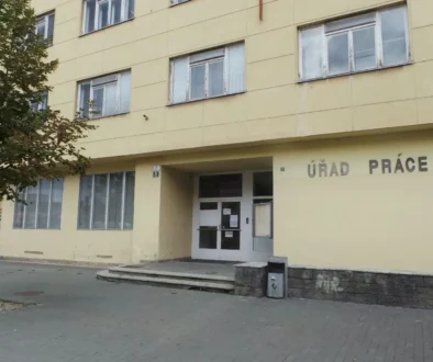 Yellow government building with the sign ÚRAD PRÁCE and glass doors at the entrance; a tree is on the left and parked cars in the background.