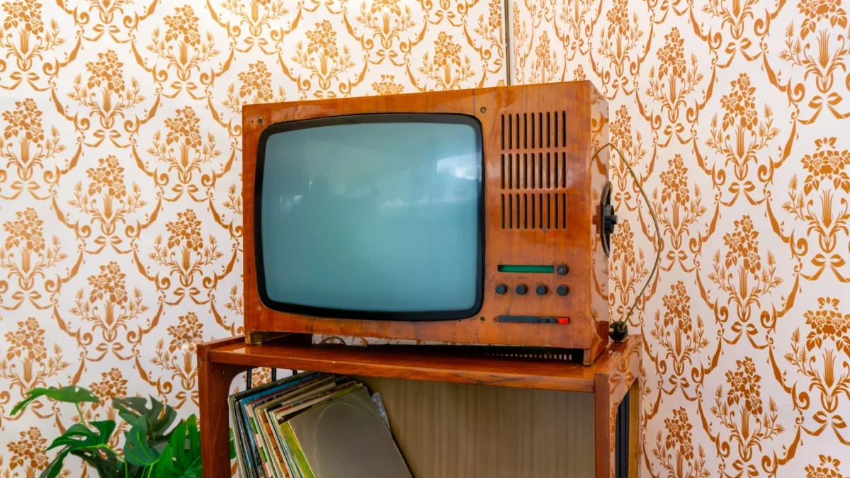 Vintage wooden TV with a curved screen on a shelf, against orange floral wallpaper and a stack of records nearby.