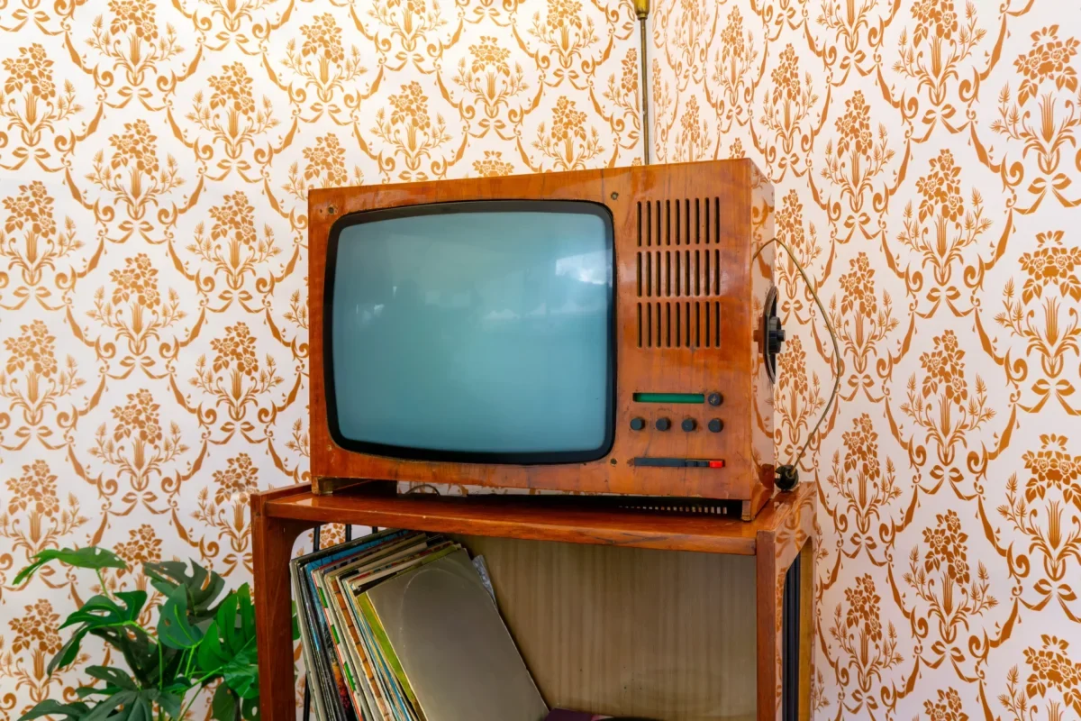 Vintage wooden TV with a curved screen on a shelf, against orange floral wallpaper and a stack of records nearby.