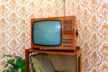 Vintage wooden TV with a curved screen on a shelf, against orange floral wallpaper and a stack of records nearby.