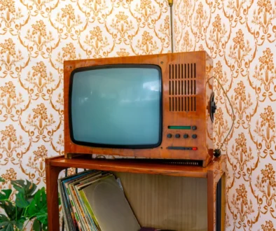 Vintage wooden TV with a curved screen on a shelf, against orange floral wallpaper and a stack of records nearby.