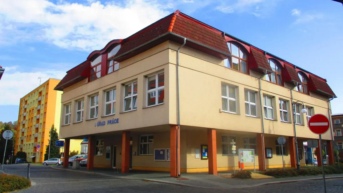 Beige multi‑story building with red tiled roof and large windows on a cobblestone street under a blue sky.