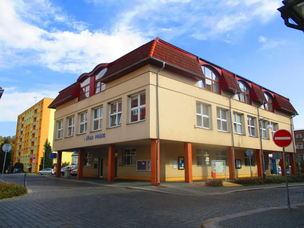 Beige multi‑story building with red tiled roof and large windows on a cobblestone street under a blue sky.