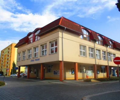 Beige multi‑story building with red tiled roof and large windows on a cobblestone street under a blue sky.