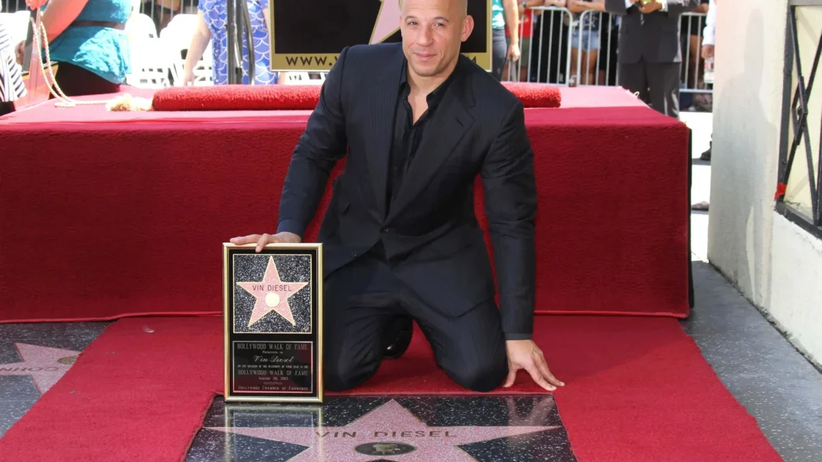 Vin Diesel kneels on a red carpet, holding his Hollywood Walk of Fame plaque in front of his star on the sidewalk.