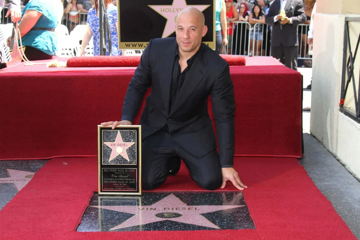 Vin Diesel kneels on a red carpet, holding his Hollywood Walk of Fame plaque in front of his star on the sidewalk.