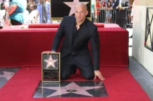 Vin Diesel kneels on a red carpet, holding his Hollywood Walk of Fame plaque in front of his star on the sidewalk.