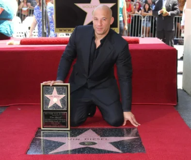 Vin Diesel kneels on a red carpet, holding his Hollywood Walk of Fame plaque in front of his star on the sidewalk.