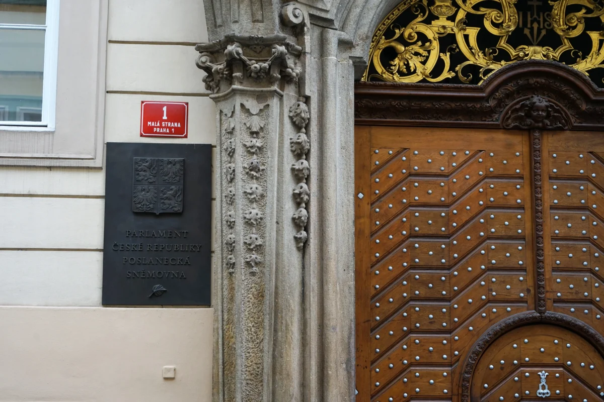 shutterstock_2173823001 1 | TVGURU.cz Historic building entrance featuring a carved stone column, red address plaque reading'1 MALÁ STRANA PRAHA 1', and a large studded wooden door beneath gilded metalwork.