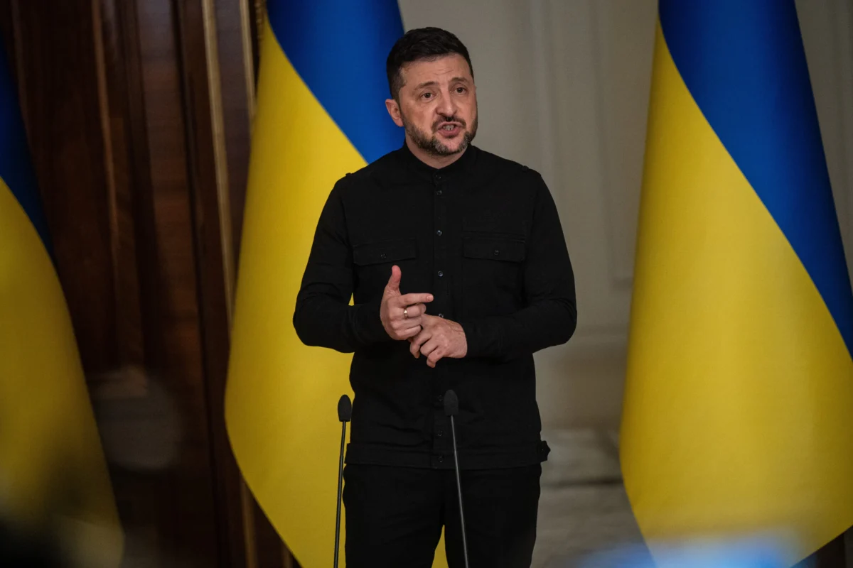 shutterstock_2751995431 1 | TVGURU.cz Man in black shirt delivering a speech, standing in front of blue-and-yellow Ukrainian flags.