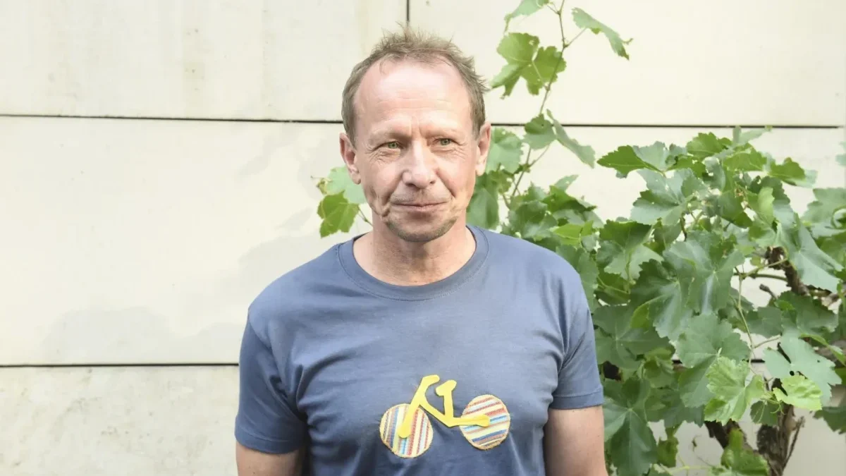 Middle-aged man in a blue t-shirt standing outdoors beside leafy vines against a pale wall.