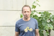 Middle-aged man in a blue t-shirt standing outdoors beside leafy vines against a pale wall.