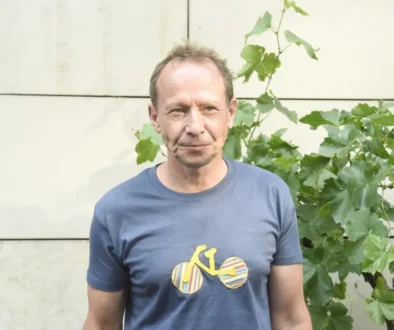 Middle-aged man in a blue t-shirt standing outdoors beside leafy vines against a pale wall.