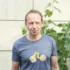 Middle-aged man in a blue t-shirt standing outdoors beside leafy vines against a pale wall.