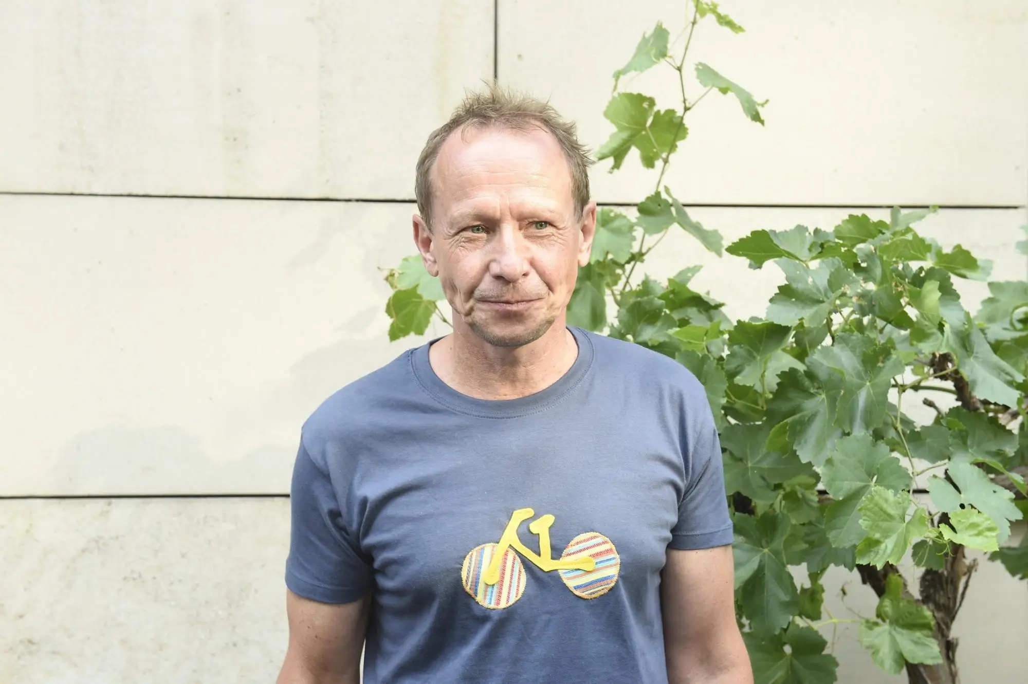 Middle-aged man in a blue t-shirt standing outdoors beside leafy vines against a pale wall.