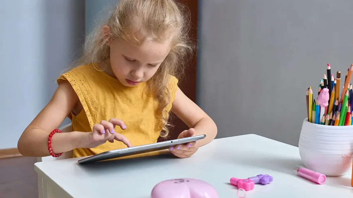 Malá holčička ve žlutém tričku sedí za stolem a dívá se do tabletu.