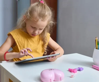 Malá holčička ve žlutém tričku sedí za stolem a dívá se do tabletu.