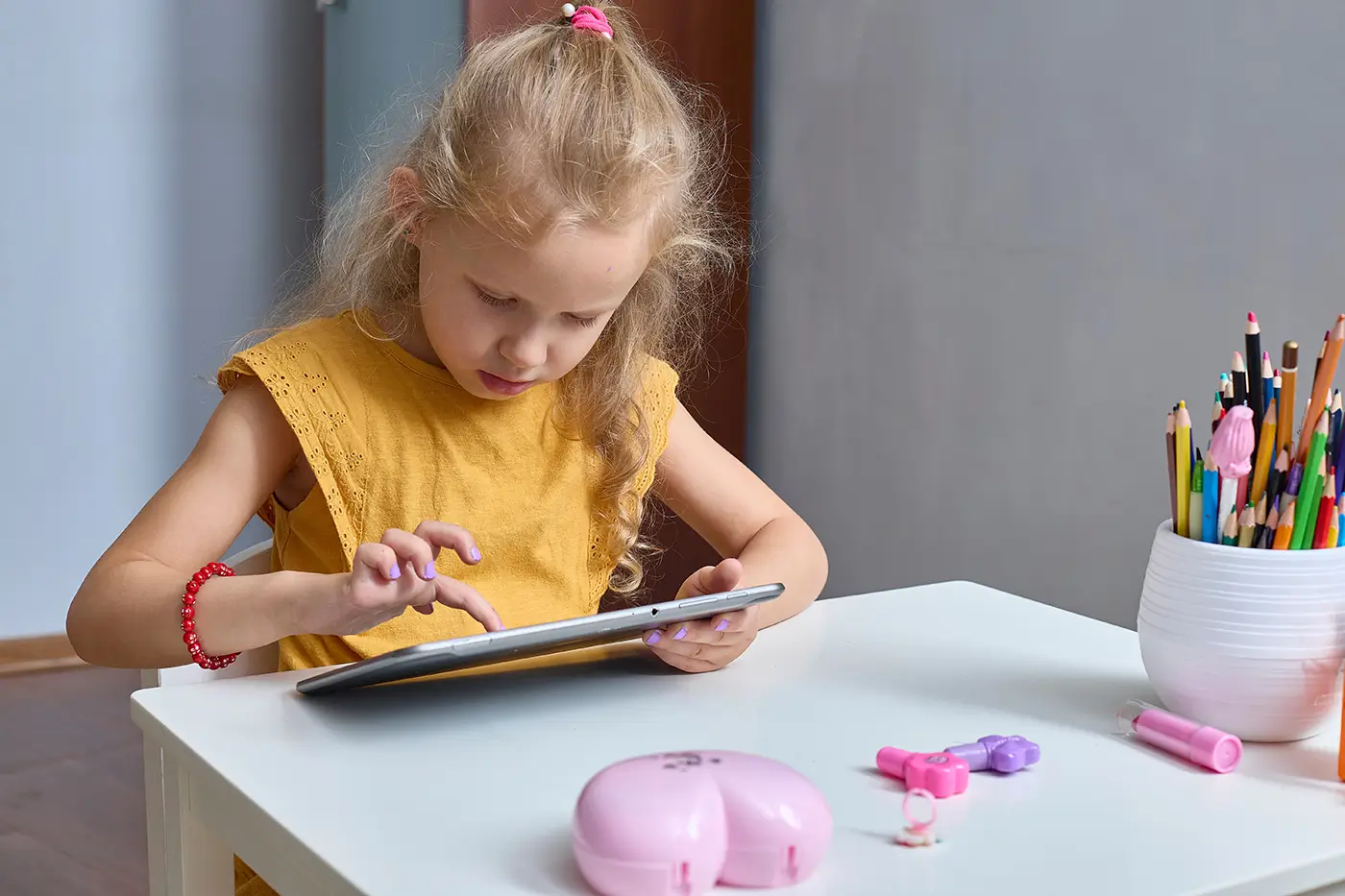 Malá holčička ve žlutém tričku sedí za stolem a dívá se do tabletu.