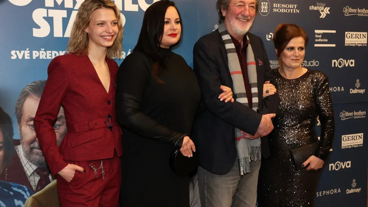 Four adults pose together for a photo at a film premiere, standing against a blue sponsorship wall; two women in red and black outfits flank a man with a scarf and a woman in a sequined dress are smiling.