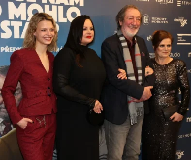 Four adults pose together for a photo at a film premiere, standing against a blue sponsorship wall; two women in red and black outfits flank a man with a scarf and a woman in a sequined dress are smiling.