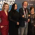 Four adults pose together for a photo at a film premiere, standing against a blue sponsorship wall; two women in red and black outfits flank a man with a scarf and a woman in a sequined dress are smiling.
