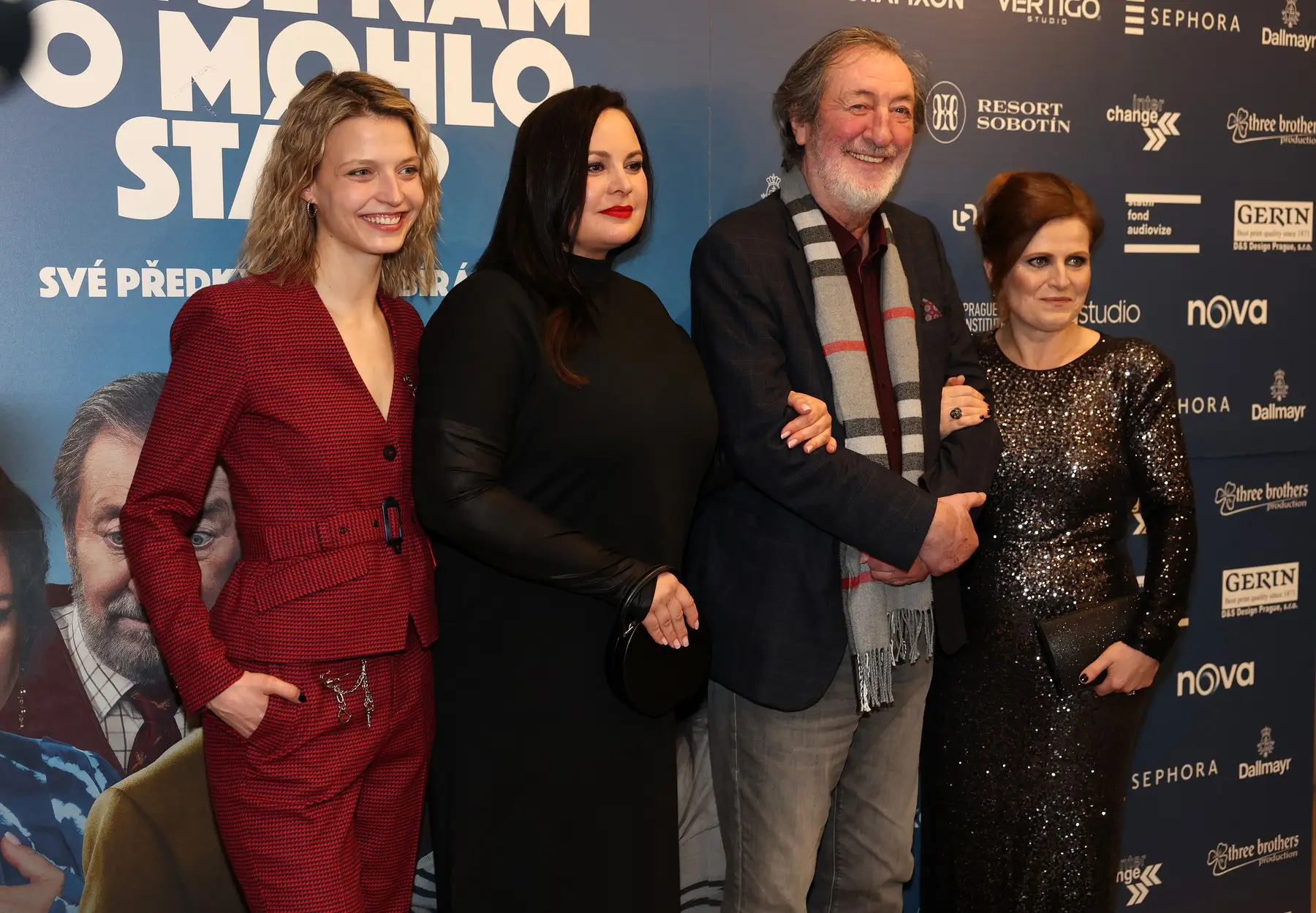 Four adults pose together for a photo at a film premiere, standing against a blue sponsorship wall; two women in red and black outfits flank a man with a scarf and a woman in a sequined dress are smiling.