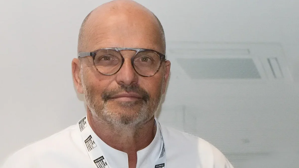 Portrait of a bald man with round glasses and light stubble, wearing a white coat and a Makro lanyard against a plain background.