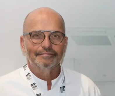 Portrait of a bald man with round glasses and light stubble, wearing a white coat and a Makro lanyard against a plain background.