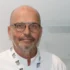 Portrait of a bald man with round glasses and light stubble, wearing a white coat and a Makro lanyard against a plain background.