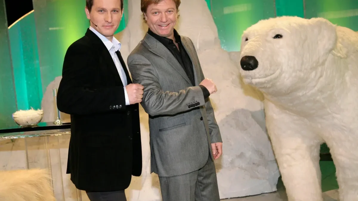 Two men in suits posing and fist-bumping beside a large white polar bear statue indoors with a green backdrop.