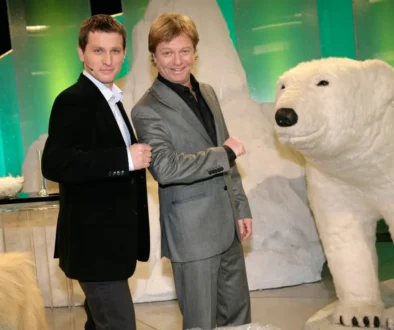 Two men in suits posing and fist-bumping beside a large white polar bear statue indoors with a green backdrop.