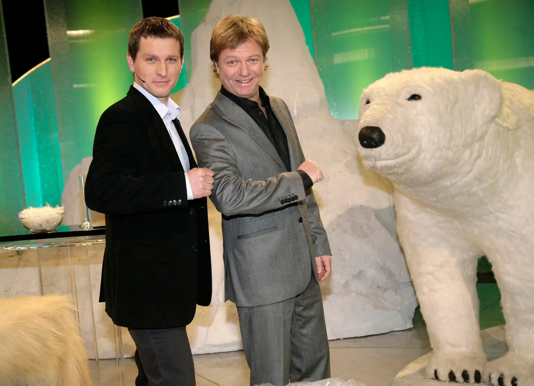 Two men in suits posing and fist-bumping beside a large white polar bear statue indoors with a green backdrop.