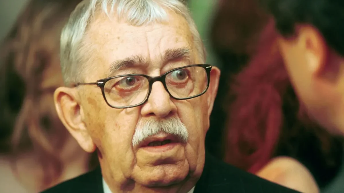 Close-up of an older man with gray hair and glasses wearing a pinstripe suit and red tie at a formal event.