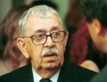 Close-up of an older man with gray hair and glasses wearing a pinstripe suit and red tie at a formal event.