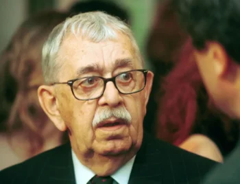 Close-up of an older man with gray hair and glasses wearing a pinstripe suit and red tie at a formal event.