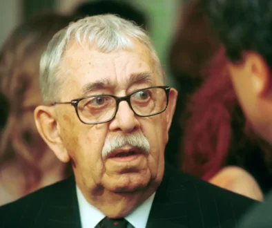 Close-up of an older man with gray hair and glasses wearing a pinstripe suit and red tie at a formal event.