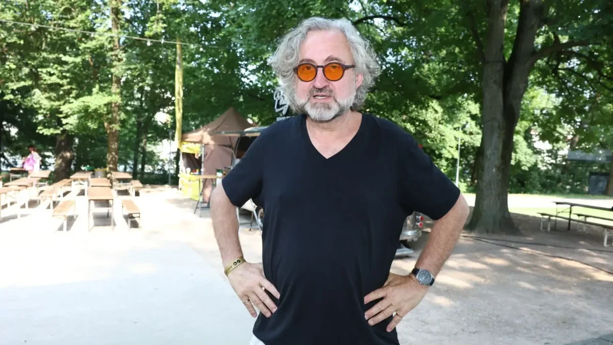 Man with gray hair and beard wearing orange round sunglasses and a black V-neck shirt standing with hands on hips outdoors, trees in the background.
