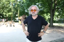 Man with gray hair and beard wearing orange round sunglasses and a black V-neck shirt standing with hands on hips outdoors, trees in the background.