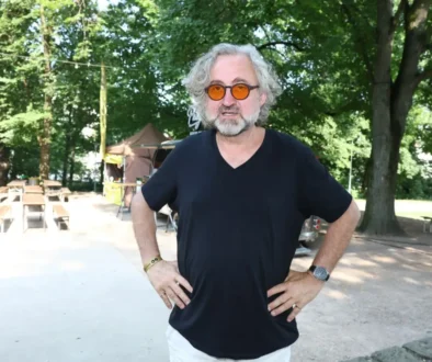 Man with gray hair and beard wearing orange round sunglasses and a black V-neck shirt standing with hands on hips outdoors, trees in the background.