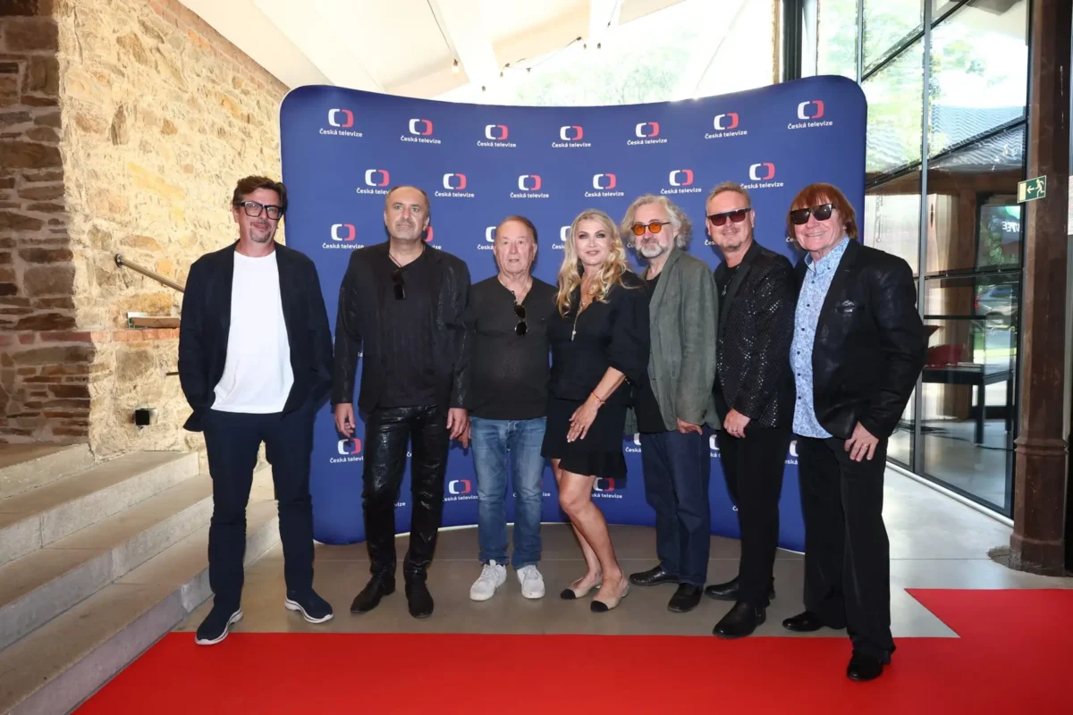 Group of eight adults posing on a red carpet in front of a blue Česká televize backdrop inside a stone-walled venue.