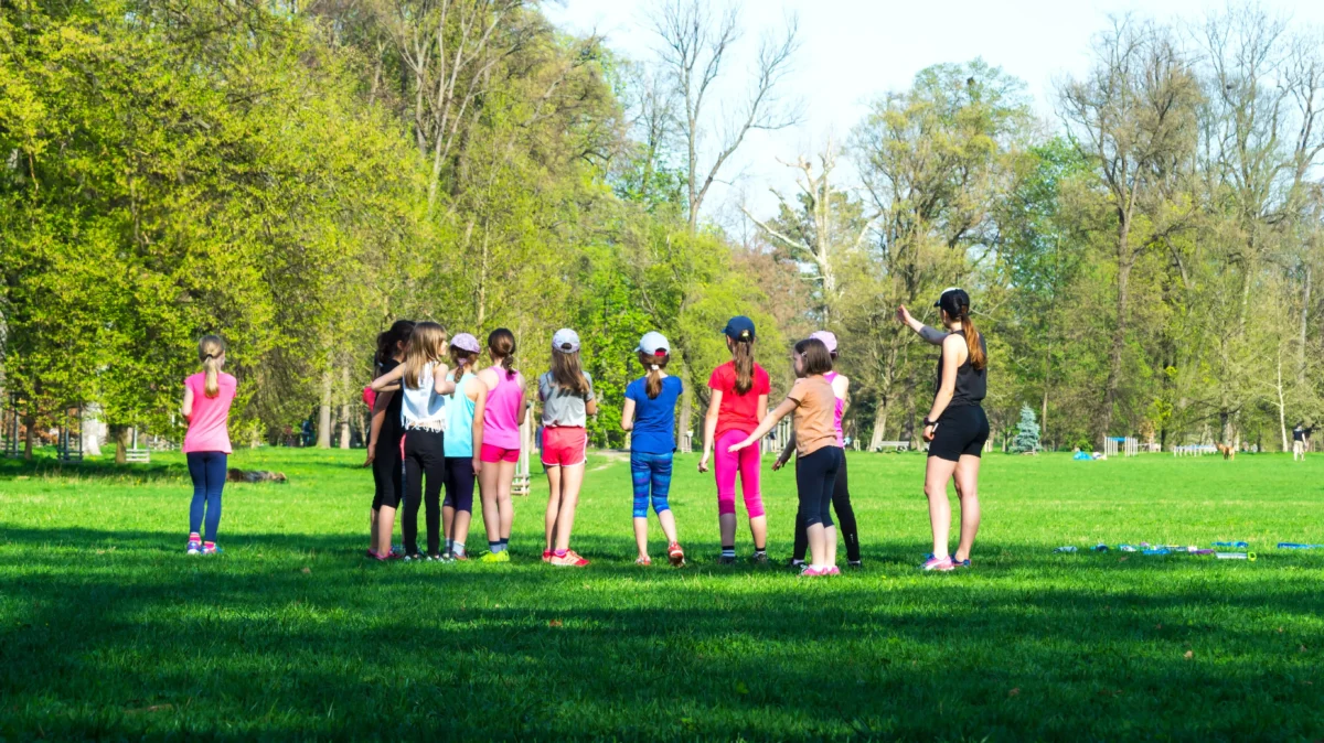Group of children in colorful athletic outfits standing in a circle in a sunny park as a coach speaks to them near trees and grassed field