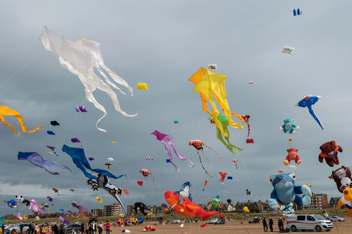 shutterstock_2518154963 1 | TVGURU.cz Dozens of colorful kites of different shapes soaring in the cloudy sky above a crowd gathered on an open field, with cars and buildings in the distance.