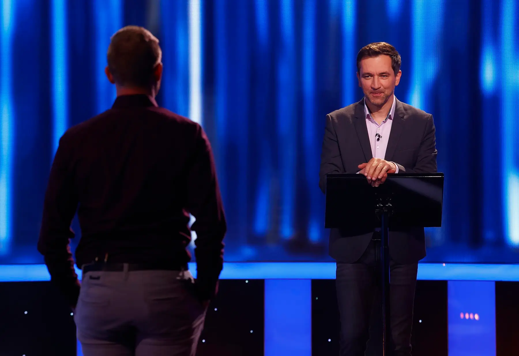 Two men on a blue-lit stage; one man faces a lectern and speaks, while the other stands with his back to the camera.